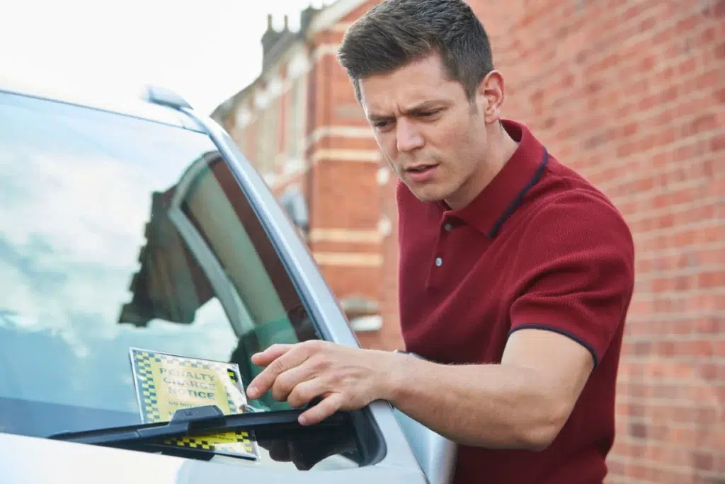 A man in a red shirt looks concerned as he examines a penalty charge notice placed on the windshield of his parked car on a city street.