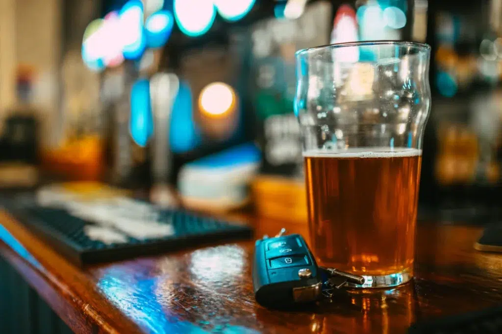 A pint of beer and a set of car keys rest on a wooden bar counter, with blurred lights and bar equipment in the background.